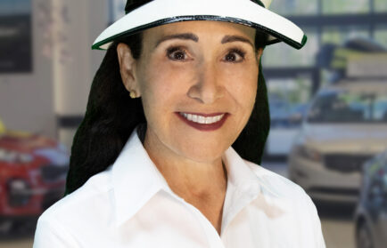 Woman wearing a white hat and white shirt with "Rick Case" logo and a name tag reading "Rita Case" in an indoor automotive showroom.