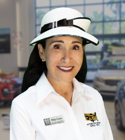 Woman wearing a white hat and white shirt with "Rick Case" logo and a name tag reading "Rita Case" in an indoor automotive showroom.