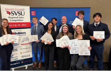 A group of nine people in business attire pose indoors, holding award certificates in front of a blue backdrop and a banner for the SVSU Regional Sales Competition.