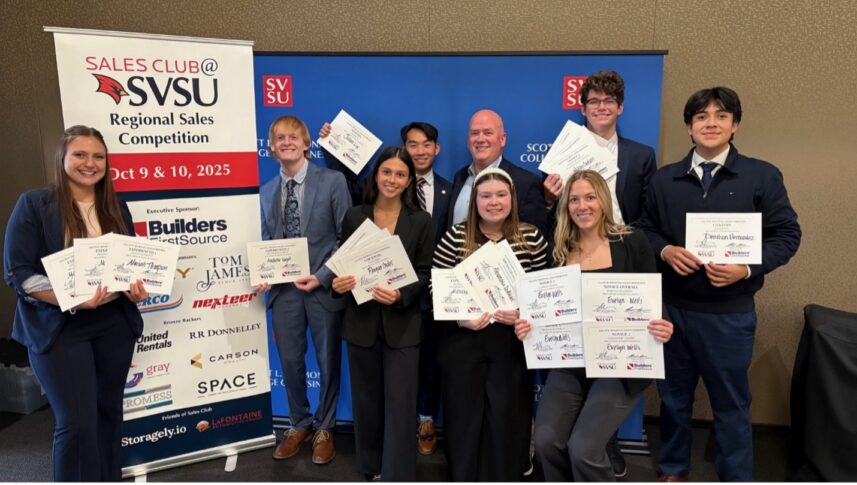 A group of nine people in business attire pose indoors, holding award certificates in front of a blue backdrop and a banner for the SVSU Regional Sales Competition.