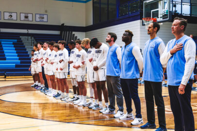 A basketball team stands in a line on a gym court, some with hands over their hearts, during a pre-game ceremony honoring the support of the Sixth Man Club.