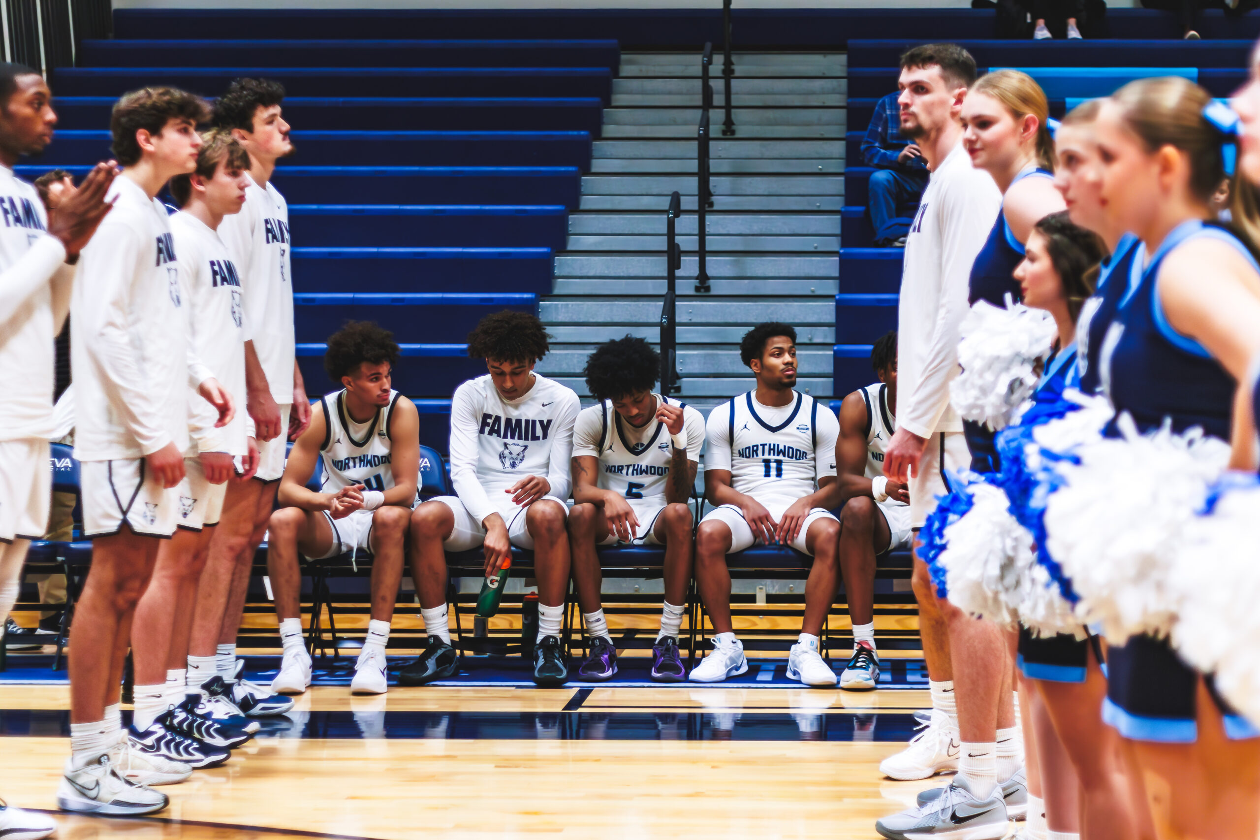Basketball players in white uniforms sit and stand on the bench, while Sixth Man Club cheerleaders in blue and white line up on the court before a game in a gymnasium.