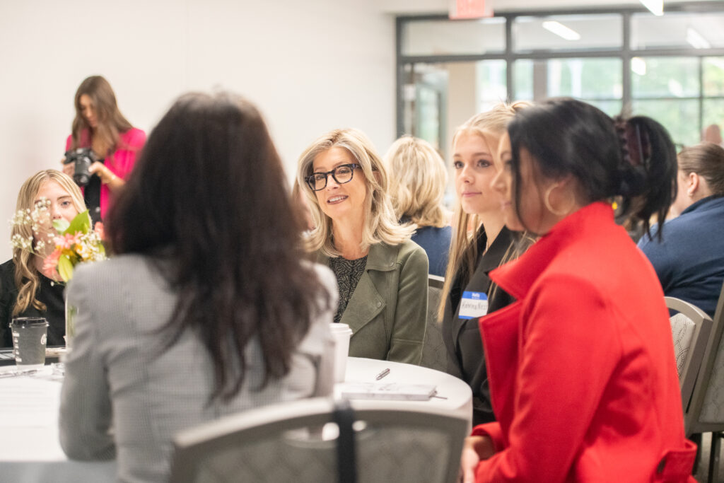 A group of women sit around a table engaged in conversation at an indoor alumni reunion; other attendees and a photographer are visible in the background.