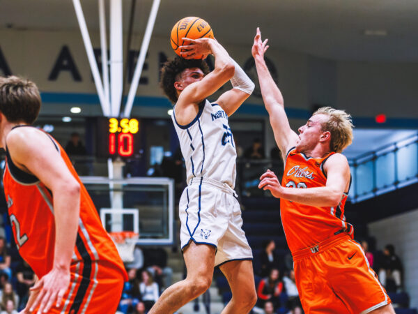 A basketball player in a white uniform jumps to take a shot while a player in orange tries to block him during a game in an indoor gymnasium, cheered on by members of the enthusiastic Sixth Man Club.