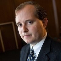 Dr. Brett M. Decker, dressed in a dark suit and patterned tie, poses for a portrait in an indoor setting with dim lighting and a blurred background.