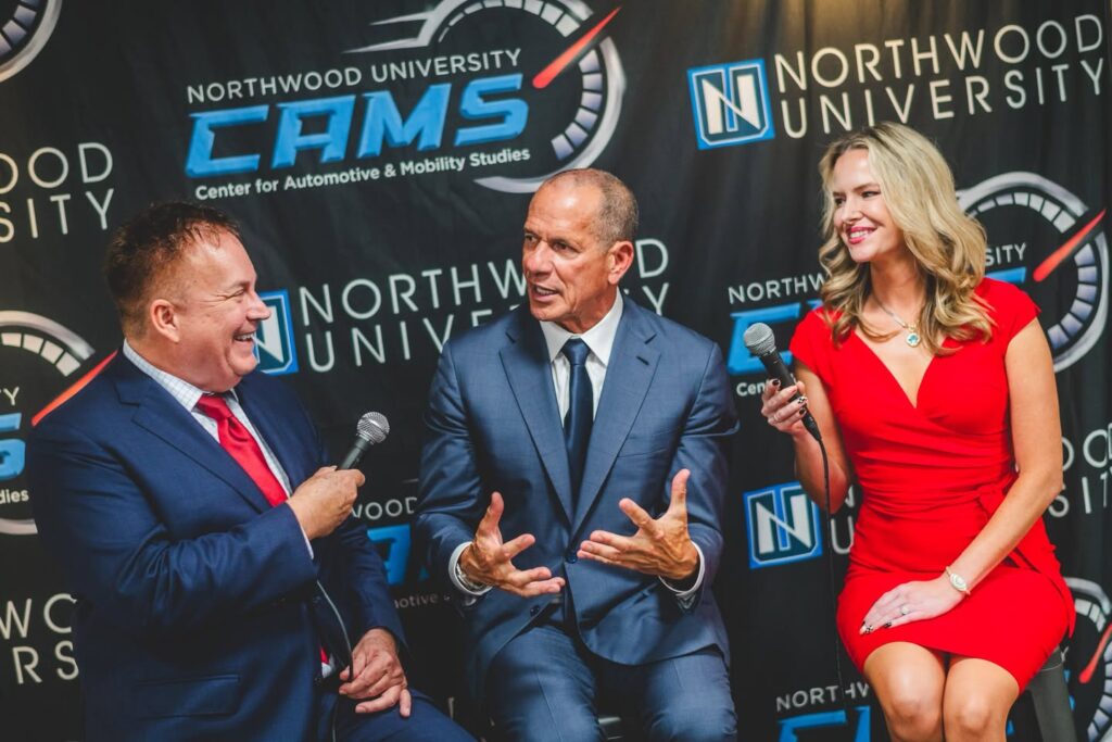 Three people in formal attire sit and talk with microphones in front of a Northwood University CAMS event backdrop during Auto Show Weekend.