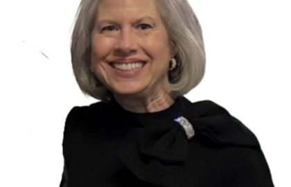 A woman with short gray hair, wearing a black top with ruffle details and a brooch, stands smiling against a plain white background, embodying the elegance celebrated by the Northwood's Distinguished Women Awards.