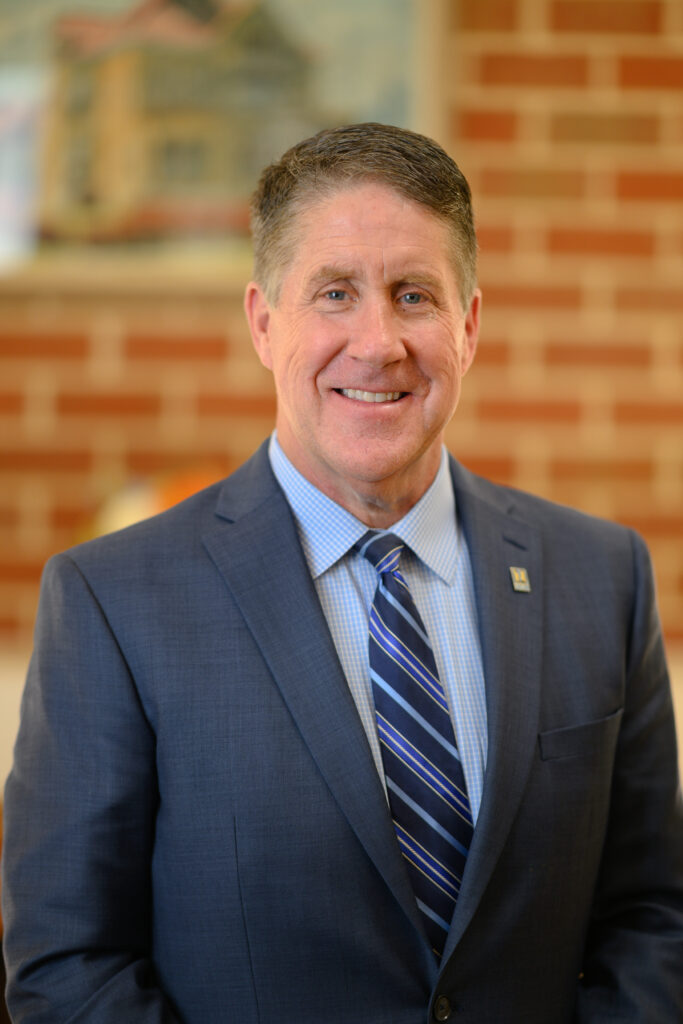 A middle-aged man in a blue suit and striped tie smiles at the camera, standing indoors with a blurred brick wall and framed artwork in the background.