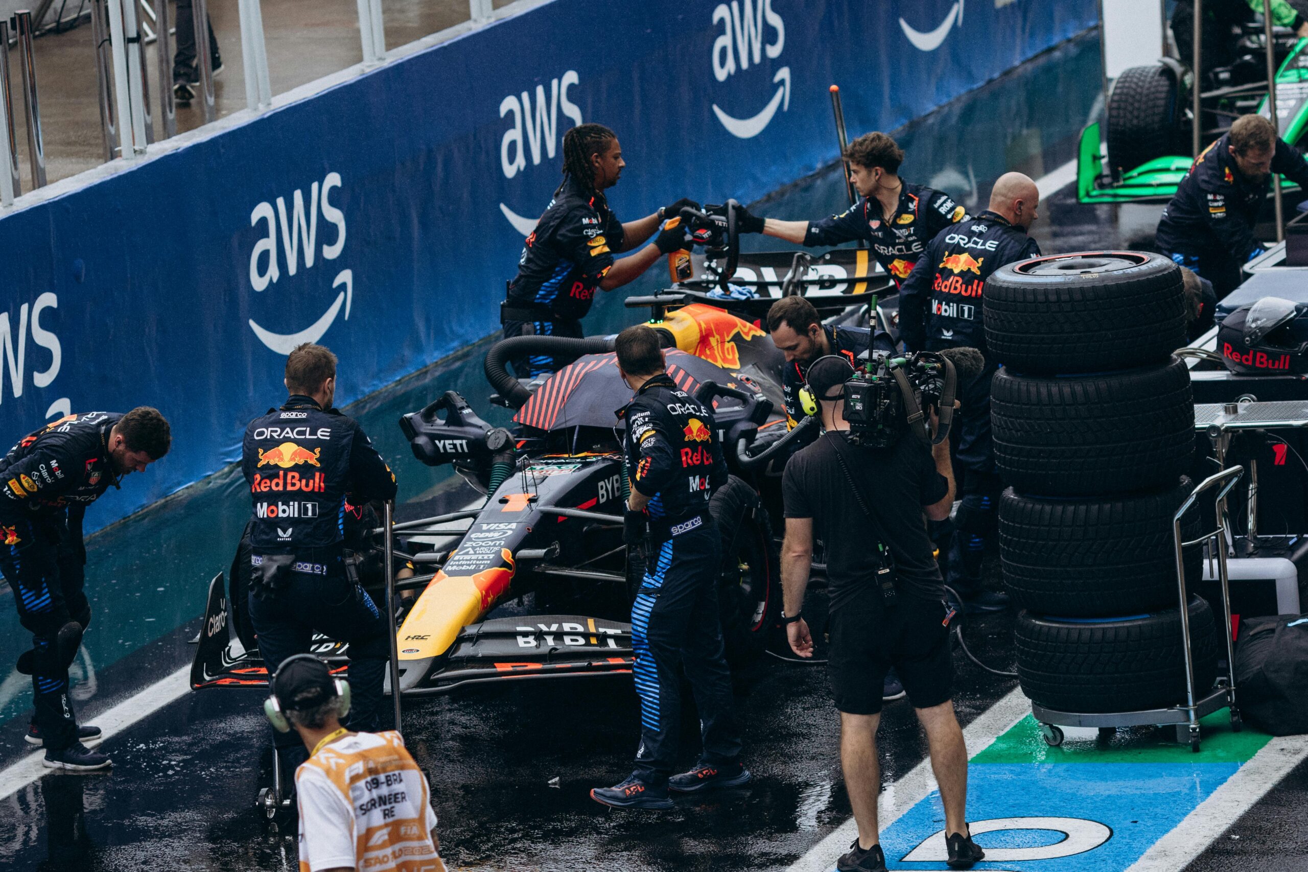 Formula 1 pit crew works on a Red Bull Racing car during a pit stop, with team members and camera crew present, stacked tires nearby—showcasing the precision and efficiency of motorsports management in high-pressure moments.