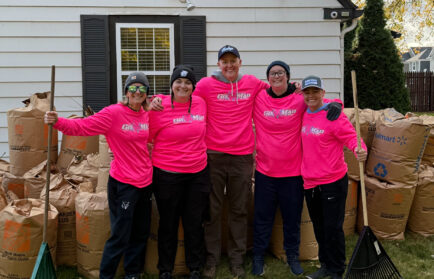Five people wearing matching pink shirts and hats stand smiling in front of a house, holding rakes, with large bags of yard waste behind them on a grassy lawn.