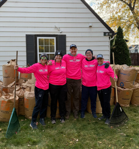 Five people wearing matching pink shirts and hats stand smiling in front of a house, holding rakes, with large bags of yard waste behind them on a grassy lawn.