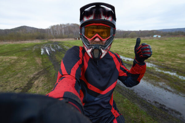 Person in red and black motocross gear and helmet gives a thumbs-up while standing on a muddy field with tire tracks in the background, showcasing the thrill and energy of motorsports.