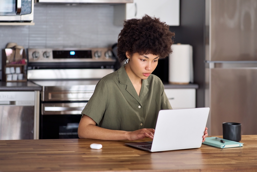 A person sits at a kitchen counter working on a laptop with a notebook, a pen, a black mug, and wireless earbuds nearby.