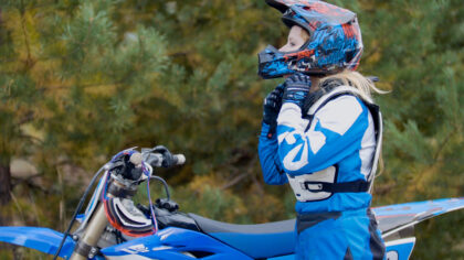 A person in blue motocross gear and helmet adjusts their chin strap while standing next to a blue dirt bike—capturing a passion for motorsports and the thrill that drives many into Motorsports Management careers.