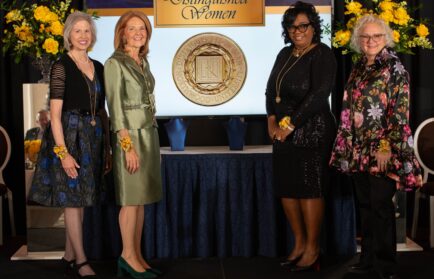 Four women stand in front of a "Distinguished Women" sign and a medallion display, all wearing formal attire and yellow floral corsages, with floral arrangements on either side.
