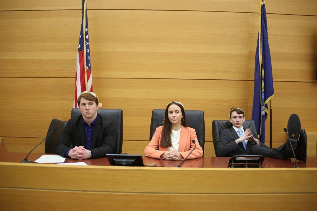 Three people in formal attire sit at a courtroom bench, with American and state flags behind them, facing forward, as part of a Mock Trial event.