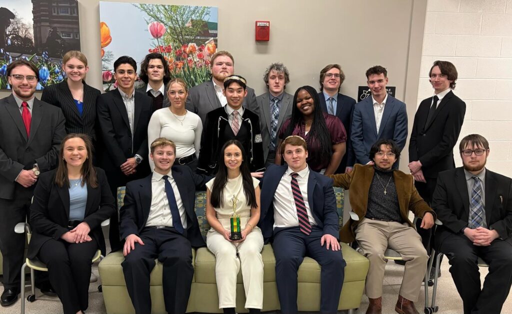 A group of eighteen people in formal attire pose indoors at a Mock Trial event, with three seated on a couch, one holding a trophy, and the rest standing behind them.