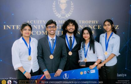Five young adults wearing medals stand together holding a trophy and a banner at the Inter University Finance Quiz Competition 2025. A backdrop displays the event name.