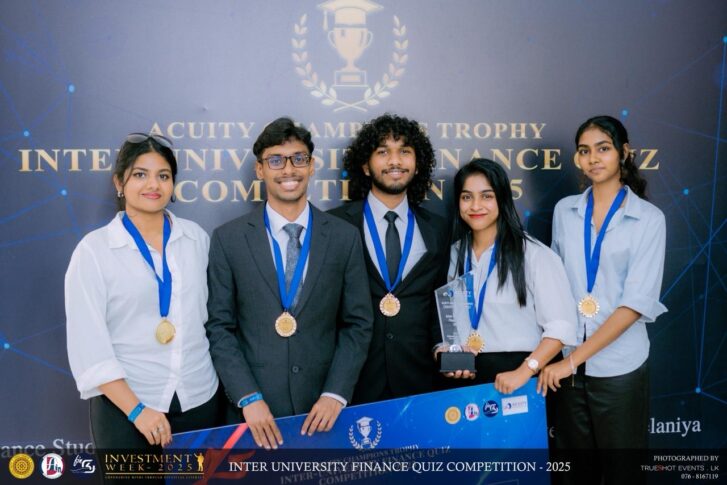 Five young adults wearing medals stand together holding a trophy and a banner at the Inter University Finance Quiz Competition 2025. A backdrop displays the event name.