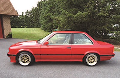 A red two-door coupe is parked on a driveway next to a lawn and trees, viewed from the side.