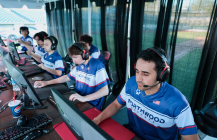 A group of young men wearing matching Northwood jerseys sit at computers with headsets, focused on playing a video game in an outdoor covered area.