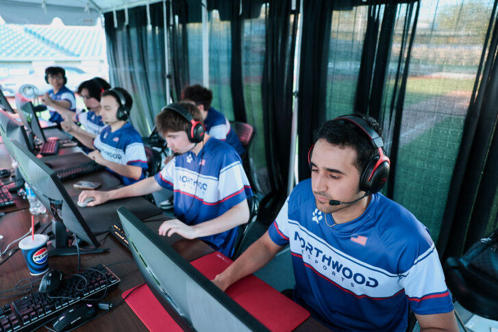 A group of young men wearing matching Northwood jerseys sit at computers with headsets, focused on playing a video game in an outdoor covered area.