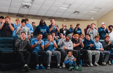 A group of young men in casual sportswear, seated in a room, smile, cheer, and clap as they watch something off-camera.