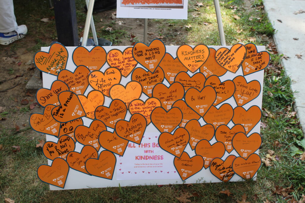 A display board outdoors is covered with orange heart-shaped notes containing handwritten messages about kindness.