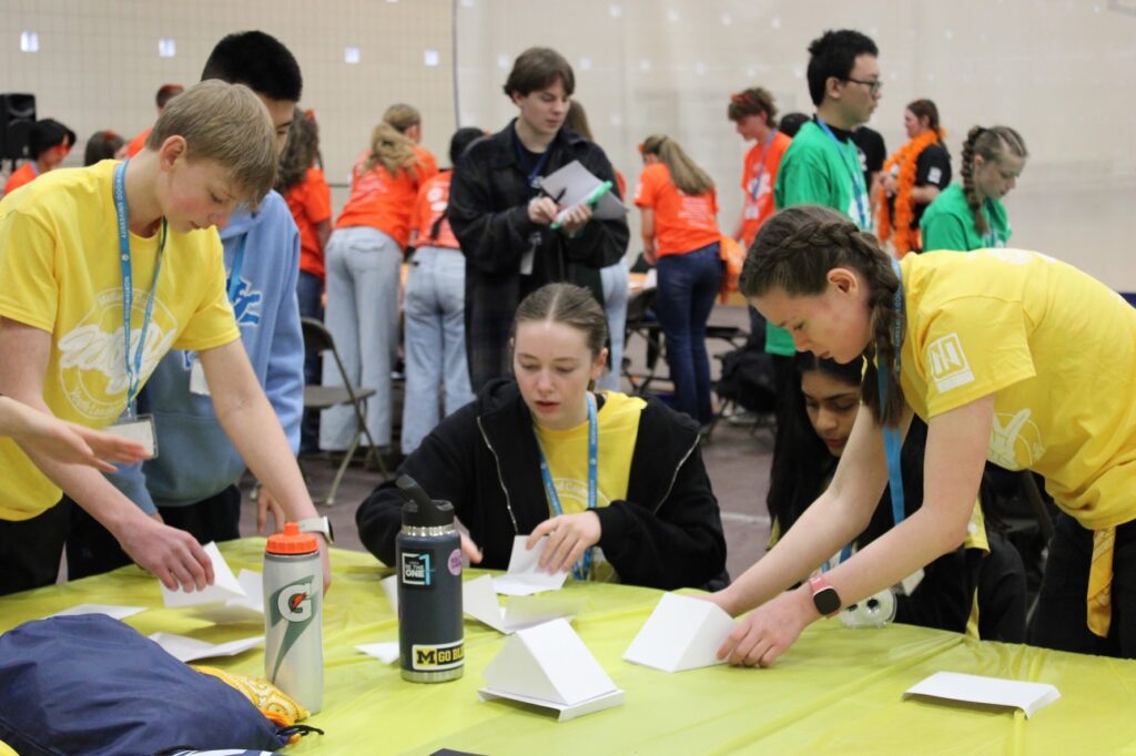 Four students in yellow shirts assemble paper structures on a table.