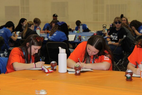 Two girls in orange shirts write on papers at a table covered with an orange tablecloth.