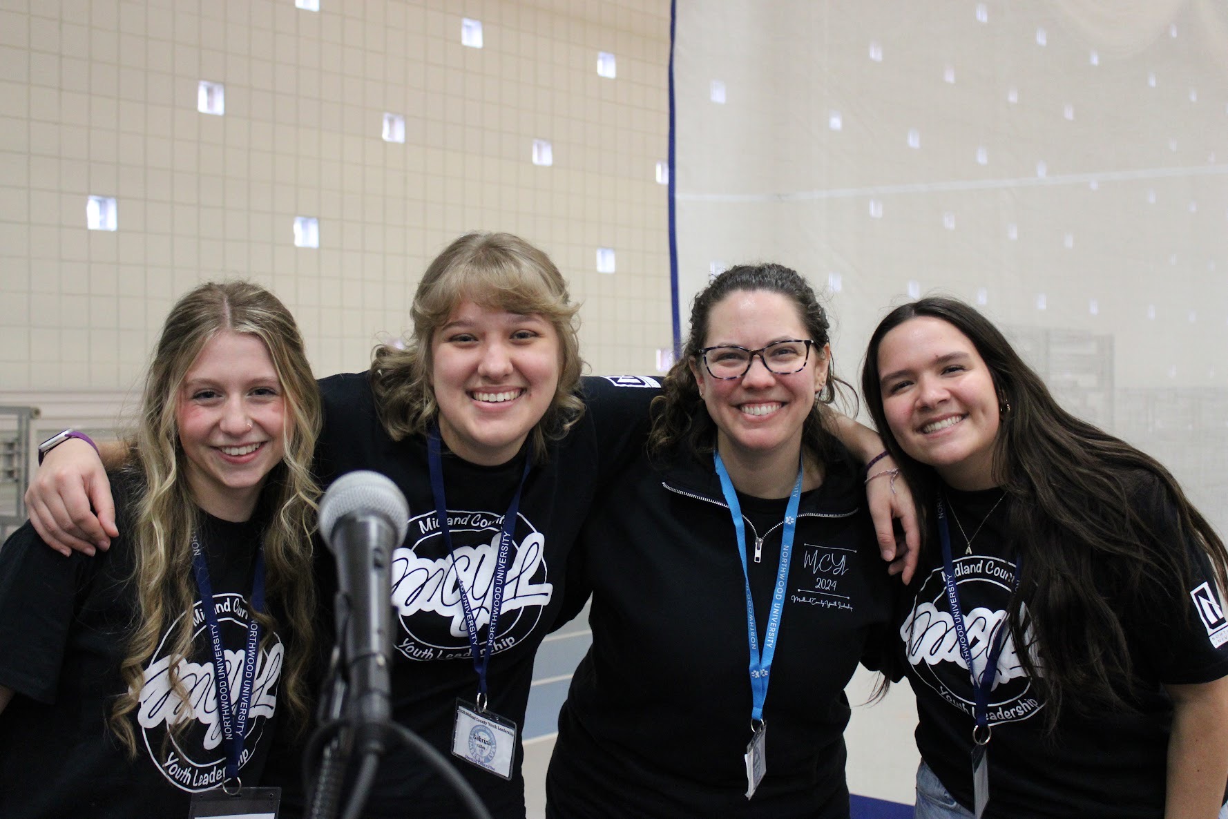 Four people stand close together smiling at the camera indoors, with one person in a black zip-up and the others in matching event t-shirts. A microphone is visible in the foreground.