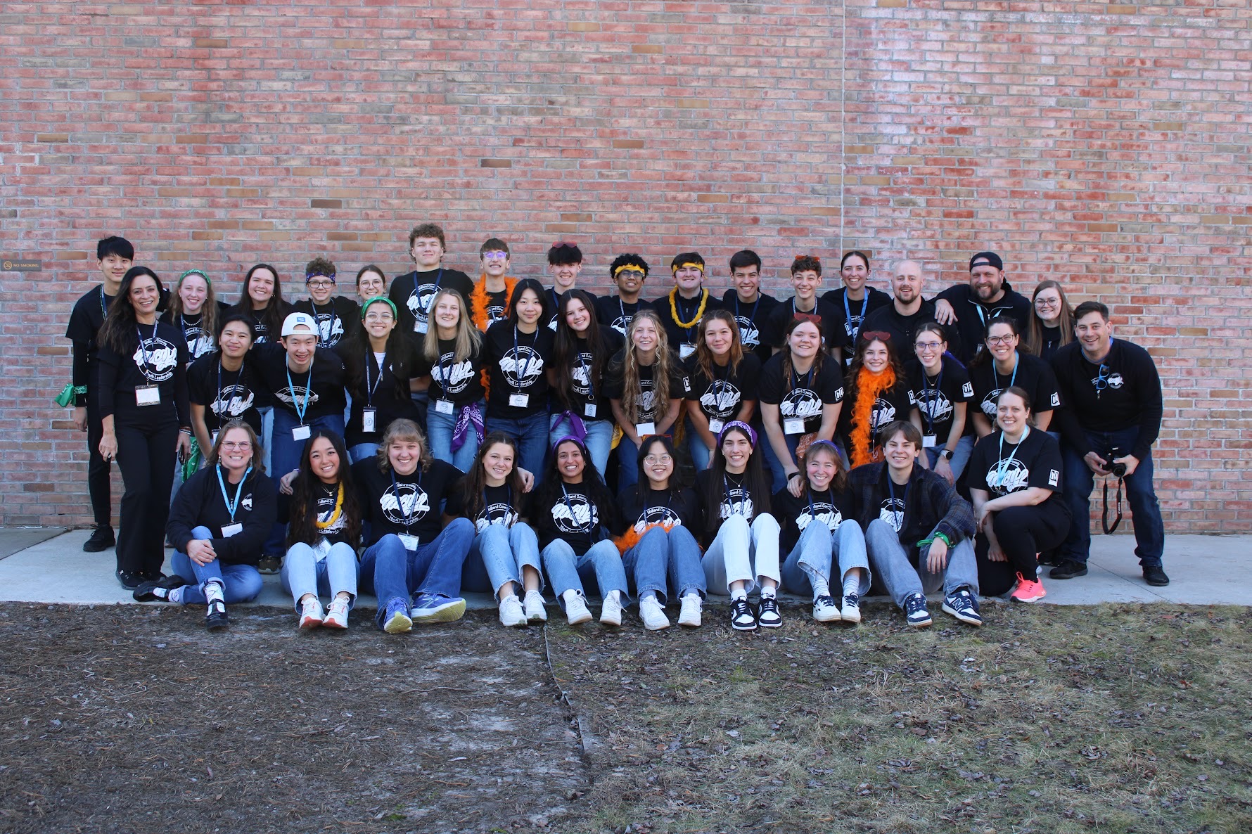 A large group of people, mostly teens, pose together in front of a brick wall, all wearing matching black shirts.