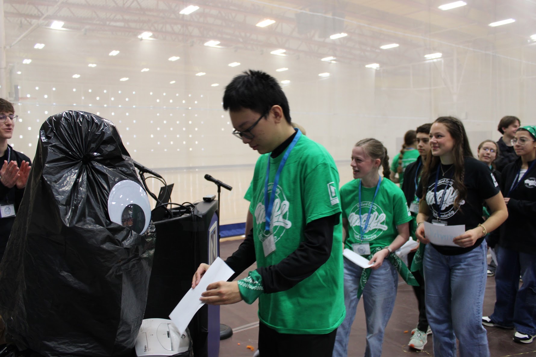 A group of students in green shirts participate in an activity, submitting papers into a box decorated as a black monster in a gymnasium setting.
