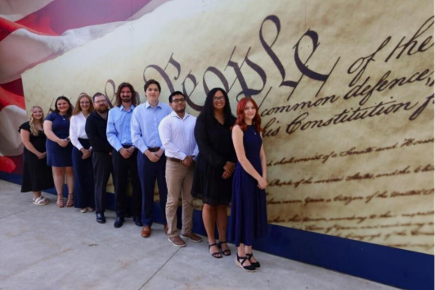 Ten people stand in a line in front of a large mural depicting the preamble of the United States Constitution at the Stafford Dinner, where a Food Network Champion was featured as keynote.