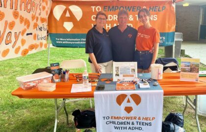 Three people stand behind a booth for a charity supporting children and teens with ADHD. The table displays brochures, pens, and promotional materials under an orange banner.