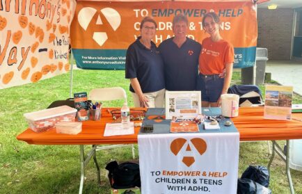 Three people stand behind a booth for a charity supporting children and teens with ADHD. The table displays brochures, pens, and promotional materials under an orange banner.
