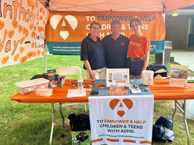 Three people stand behind a booth for a charity supporting children and teens with ADHD. The table displays brochures, pens, and promotional materials under an orange banner.
