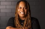 Food Network Champion with long, light brown dreadlocks and a black t-shirt smiles in front of a dark tiled wall.