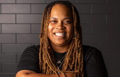 Food Network Champion with long, light brown dreadlocks and a black t-shirt smiles in front of a dark tiled wall.