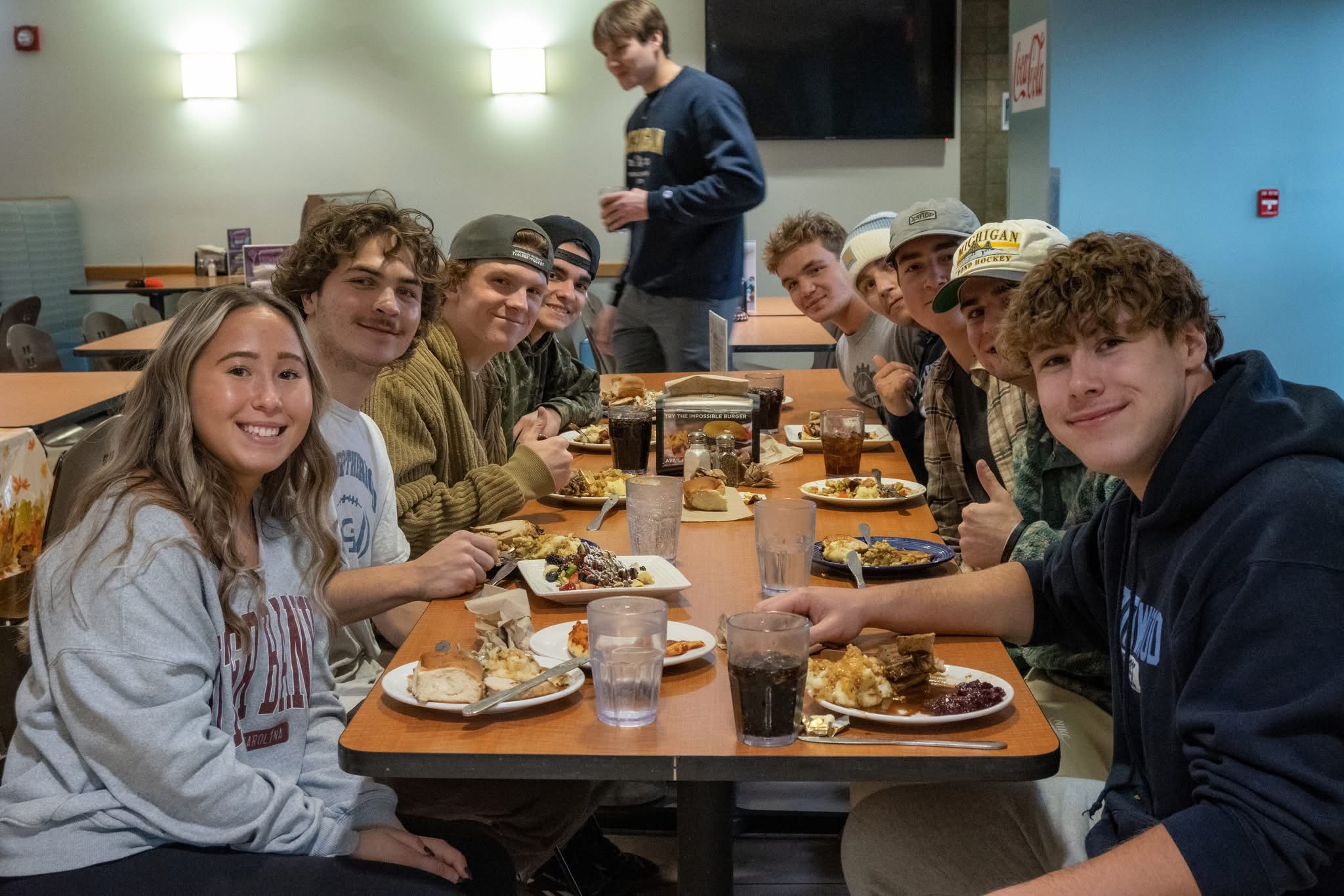 A group of young adults sit around a cafeteria table, smiling and enjoying a meal together provided by Dining Services. Plates of food and drinks are on the table, while one person stands in the background.