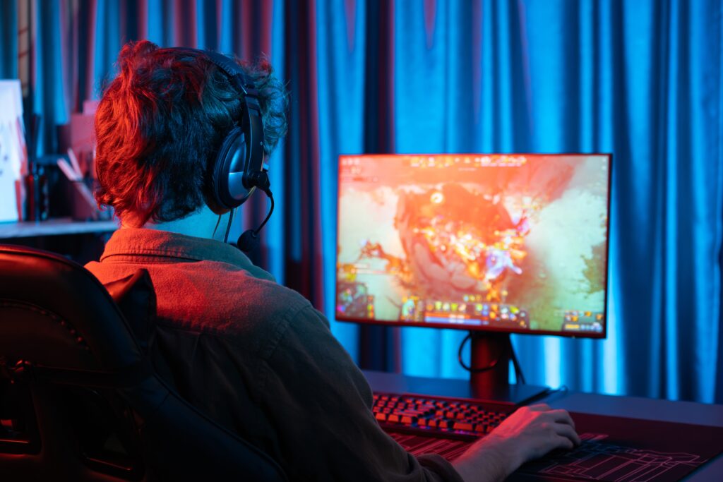 A person wearing headphones sits at a desk playing a colorful video game on a monitor in a dimly lit room with blue curtains, likely unwinding after studying for their sports management degree.