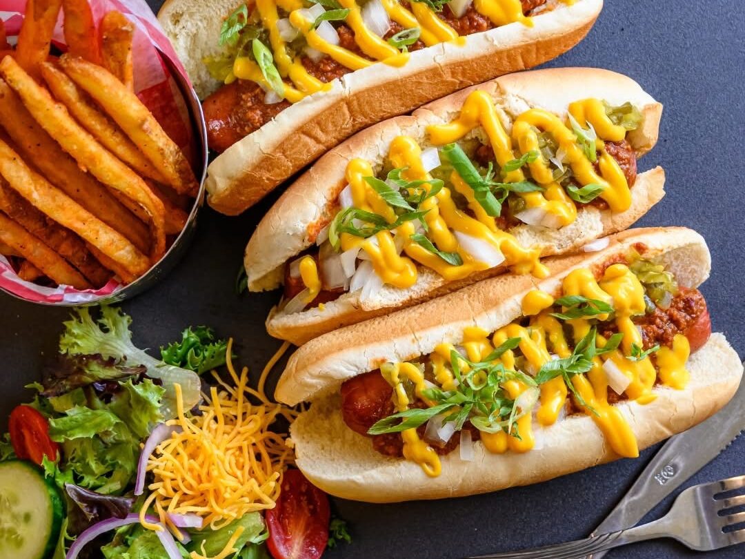 Enjoy three hot dogs topped with mustard, onions, and relish, served by Dining Services on a plate with curly fries, a side salad, shredded cheese, and a refreshing glass of soda.