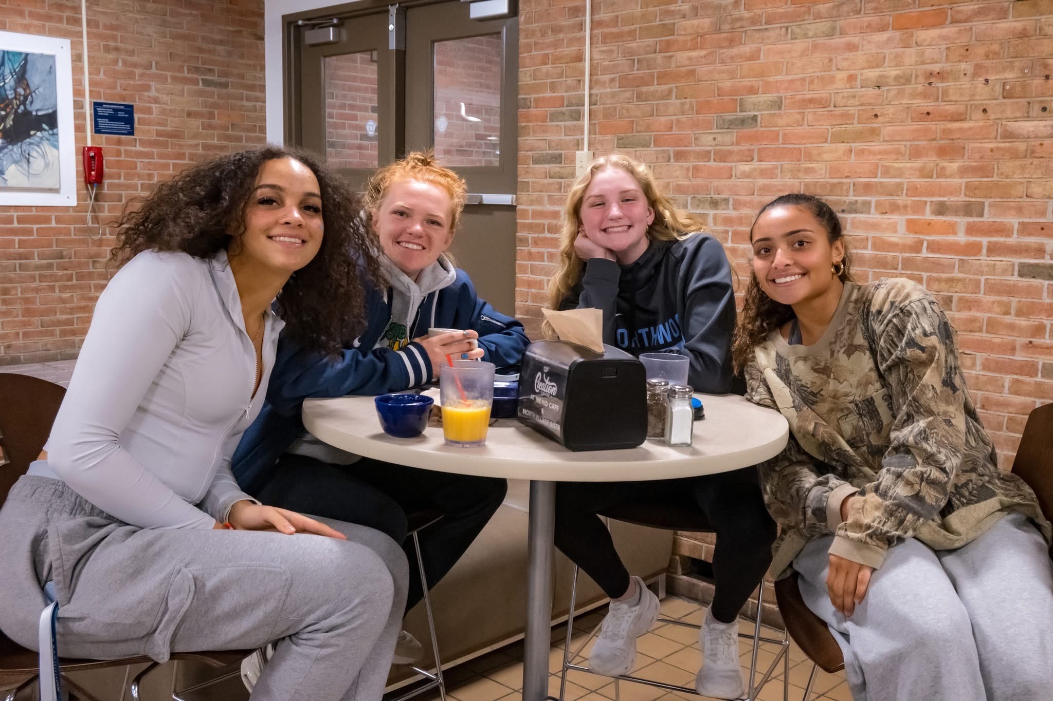 Four young women sit around a table in a cafeteria, smiling at the camera. Drinks and napkins from Dining Services are on the table, with a brick wall in the background.