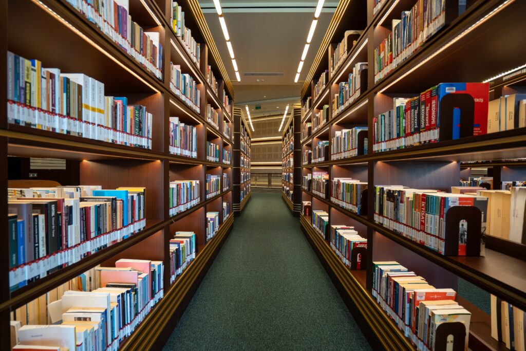 A well-lit library aisle with tall bookshelves on both sides, filled with various books to support your bachelor degree studies, and a carpeted floor leading toward the back of the library.