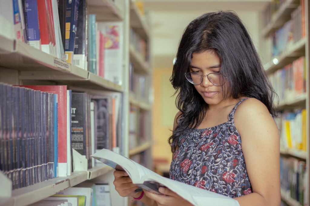 A person with long dark hair and glasses, likely pursuing a bachelor degree, reads a book while standing between bookshelves in a library.