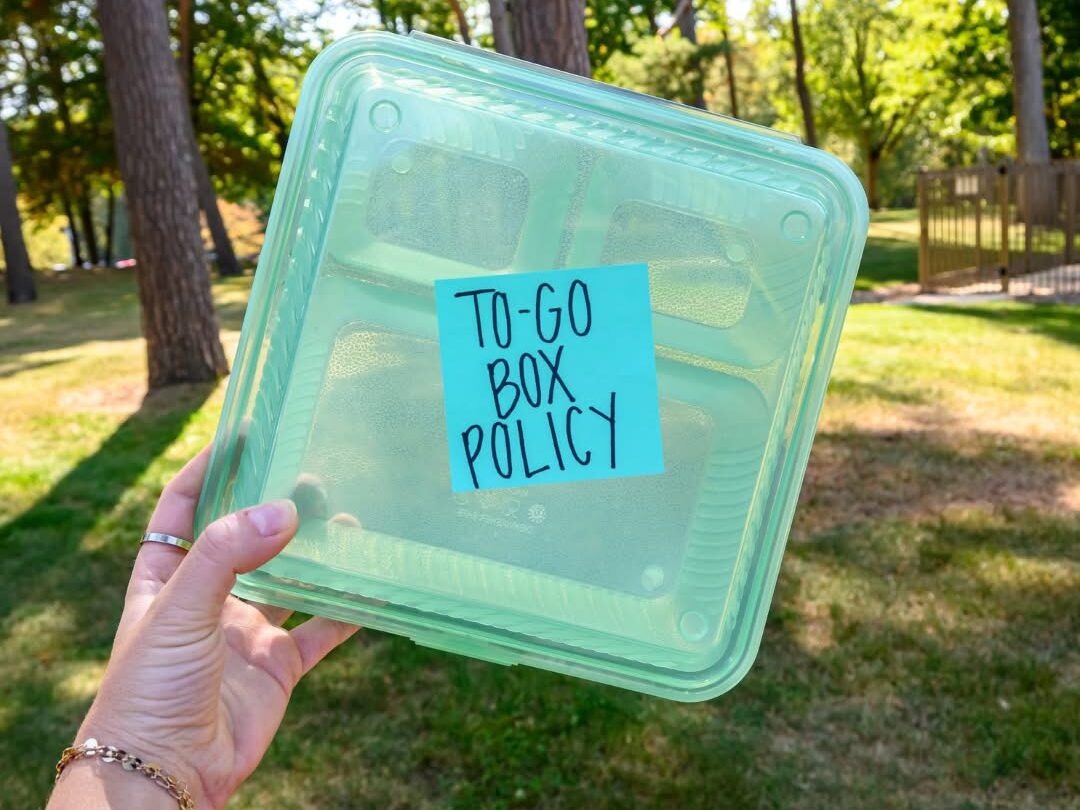 A hand holds a green plastic to-go box with a blue note labeled "TO-GO BOX POLICY" from Dining Services in an outdoor park setting.