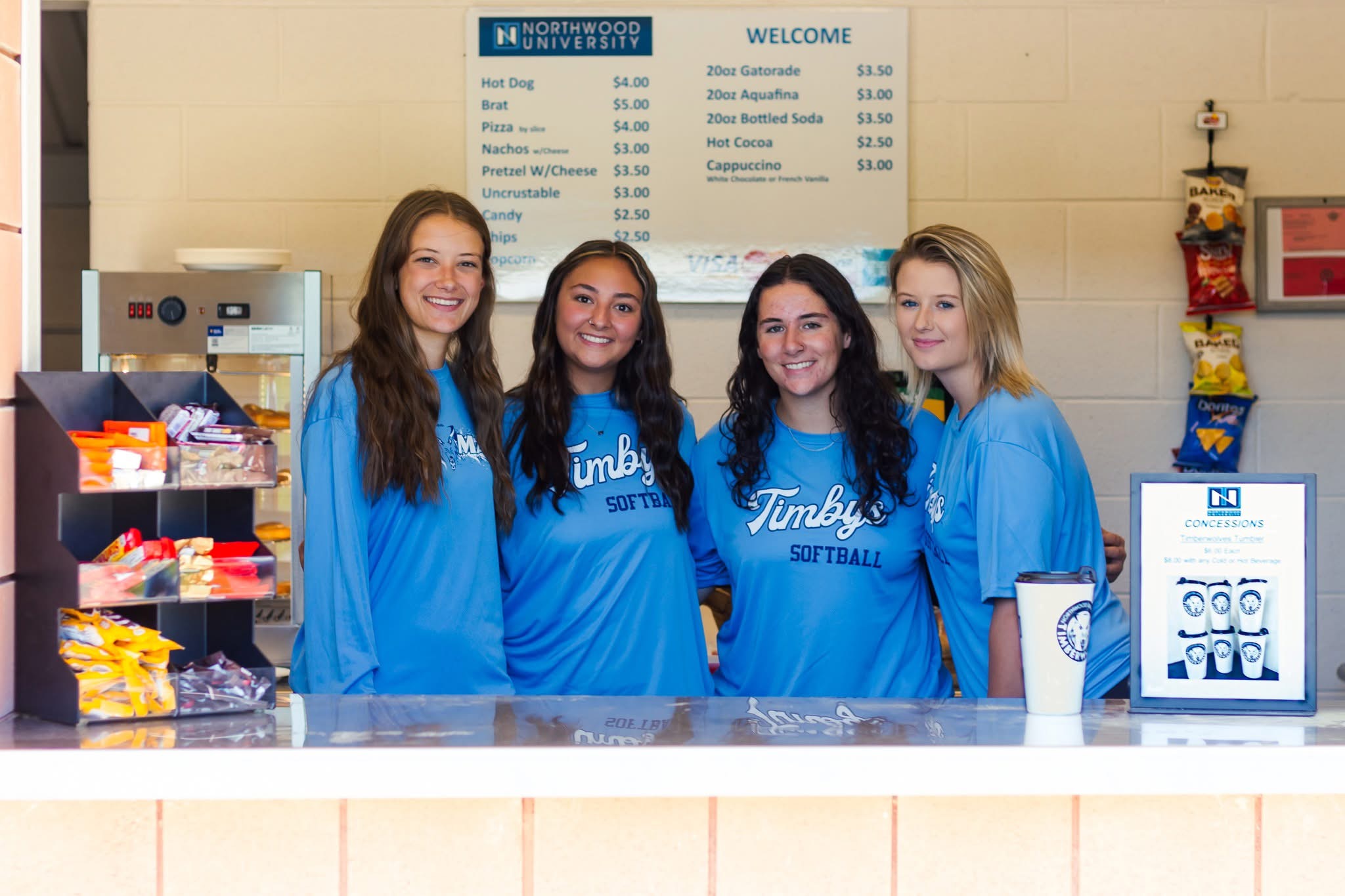 Four young women in matching blue softball shirts stand behind a concession stand counter, representing Dining Services, with snacks and a menu visible in the background.