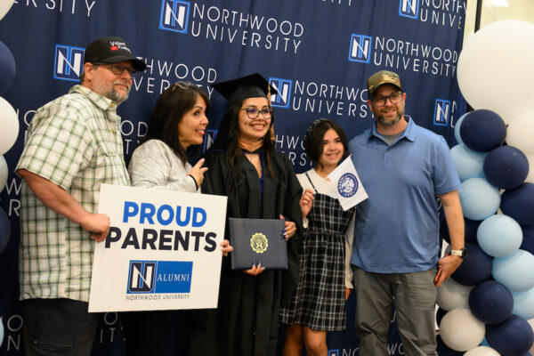 A graduate in cap and gown holds her diploma and poses with four people, including proud parents. Against a Northwood University backdrop with blue balloons, they celebrate the college benefits of Legacy students.