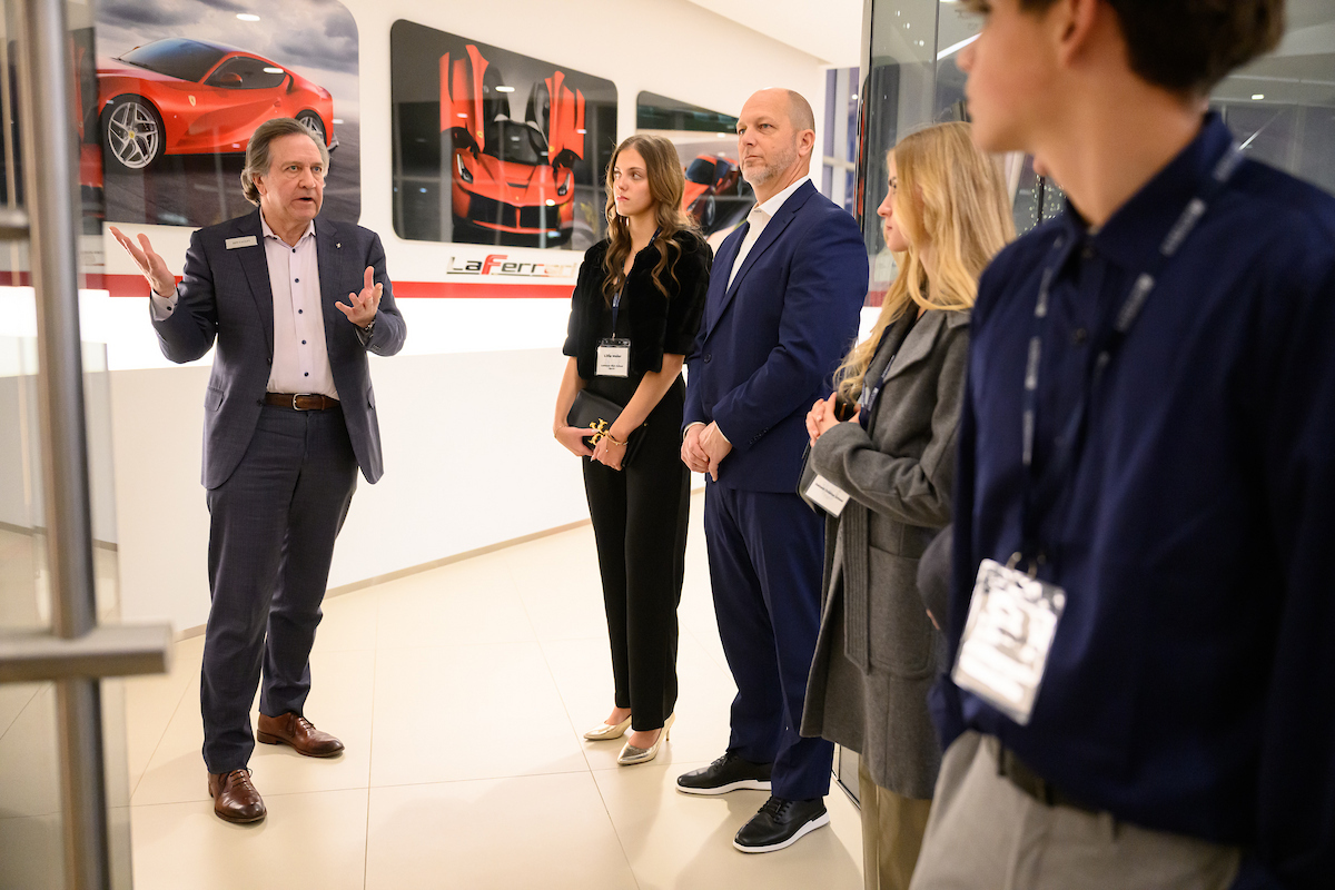 A man in a suit discusses automotive trends with four people in a modern showroom adorned with sports car photos, reflecting themes of Mobility Studies.