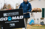 A person in a blue jacket and hat rakes leaves in a yard. A "Northwood University GO MAD" sign is displayed in the foreground.